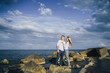 © Daria - Long shot. Attractive couple in bright clothes standing on stones in the sea against a blue cloudy sky. Lit by the soft evening sun. Looking forward, hug, kissing and enjoying beautiful evening