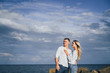 © Daria - Long shot. Attractive couple in bright clothes standing on stones in the sea against a blue cloudy sky. Lit by the soft evening sun. Looking forward, hug, kissing and enjoying beautiful evening