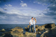 © Daria - Long shot. Attractive couple in bright clothes standing on stones in the sea against a blue cloudy sky. Lit by the soft evening sun. Looking forward, hug, kissing and enjoying beautiful evening