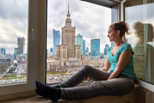 Side View Of Young Woman Sitting On Window Windowsill With Aerial High Angle View Of Warsaw, Poland Cityscape Downtown With Palace Of Culture And Science Skycrapers