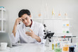 © mrwinn - A young male scientist with black hair wearing white coat looking at a mobile phone device in his hand with another hand on the head. Looking tired and stressed in a laboratory setting.