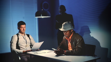 Wall Mural - Multiracial desperate criminal writing last message on smartphone in interrogation room. Police detective pulling the phone from the accused man.