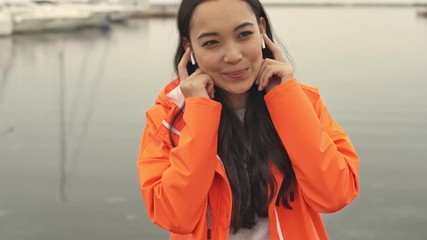 Wall Mural - Beautiful asian positive optimistic woman walking by harbor grant outdoors in orange coat listening music by wireless earphones.