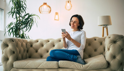 Cozy atmosphere. Beautiful woman in casual clothes is holding a smartphone in her hands and smiles while sitting on a soft couch in a designer flat.