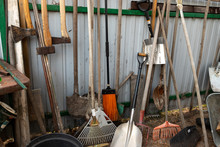 Set Of Many Different Old Rusty Vintage Gardening Equipment Near Fence At Shed Storage In House Backyard. Rustic Agricultural Home Tools At Barn Wall At Countryside Cottage