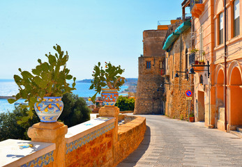 Wall Mural - Old narrow street with Ceramic pot with cactus on wall and view on the port and sea in front of Sciacca, Sicily, Italy