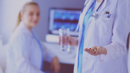 Wall Mural - shot of doctor holding pills and glass of water at clinic with colleague on the background.