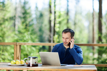 serious middle aged man talking on cell phone and using laptop computer sitting at wooden table on c