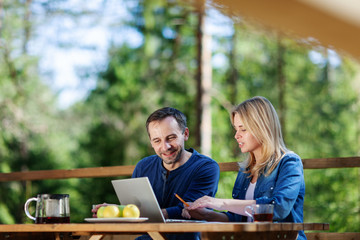 Couple sitting at wooden table on country house balcony, looking at laptop screen and discussing project in nature