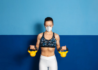 Young fit woman with n96 face mask  doing biceps exersice at the gym with kettlebells. Fitness strength workout under coronavirus health crisis.