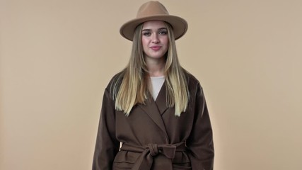 Sticker - A confident smiling young woman wearing hat and coat is showing a thumb up gesture isolated over the beige background in studio