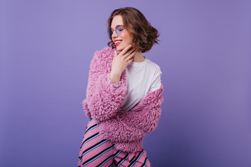 Wall Mural - Smiling white girl with short haircut posing on purple background. Studio shot of graceful european lady in bright fluffy jacket.