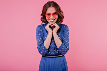 Wall Mural - Romantic brunette lady in elegant blue attire posing in studio with smile. Indoor shot of beautiful female model in sunglasses laughing on pink background.