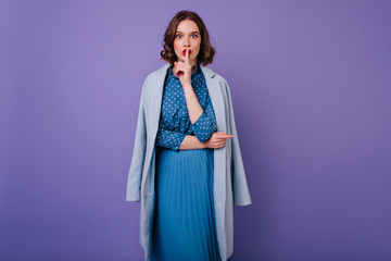 Attractive girl in vintage dress posing in studio with purple background. Indoor photo of inspired curly lady in blue coat wants silence.