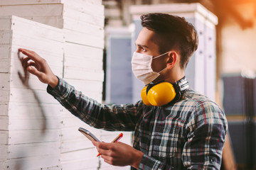 Wall Mural - Portrait male foreman inspector in medical face mask and protective headphones checking woodwork stock at factory storage. Man supervisor counting wood inventory. Warehouse worker. COVID-19 quarantine