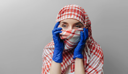 Muslim woman in keffiyeh, protective medical protect mask and blue latex gloves on a gray background. The concept of quarantine, coronavirus, pandemic COVID-19. Copy space.