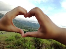 Cropped Hands Making Heart Shape On Grassy Field Against Cloudy Sky