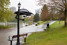 Light Pole Constructed With Arms Holding Umbrella In Front Of Lake On Rainy Day. Walking Path In Background.