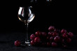 © Adria - a glass glass stands on the table against a black background. There are red grapes nearby. On top of the glass is poured wine from a bottle with a dark glass