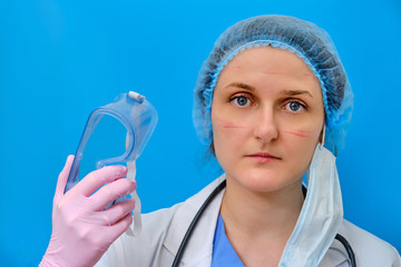 A doctor with a wounded face removes goggles after working in the hospital, blue background. A nurse in a white coat and a medical beret with marks on her face. Concept stay home, coronavirus.