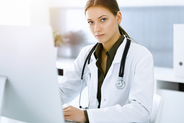 Woman-doctor typing on pc computer while sitting at the desk in sunny hospital office. Data in medicine and healthcare