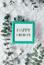 Text "Happy Holidays" In Vibrant Green Frame. Christmas, New Year Winter Frame With Fresh Eucalyptus Twigs And White Black Trinkets, Baubles With Stars. Flat Lay, Top View On White Marble Background.