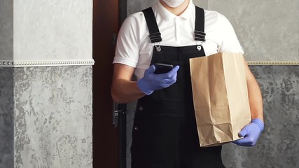 Wall Mural - courier in surgical mask medical gloves with online food from grocery in paper bag. delivery man at doorway delivering online order and check phone. contact free less delivery service in quarantine