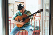 © ANTONIO HUGO - Bohemian musician with mask on his face and hat playing the Spanish guitar from his balcony. Dressed in a Hawaiian shirt and jeans Concept music from home during confinement. Coronavirus pandemic.