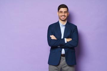 Modern businessman dressed in blue smart casual suit, wearing trendy glasses, standing with arms crossed, isolated on purple background, copyspace on left