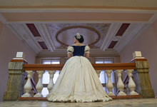 Beautiful Girl, In A Romantic Dress, Retro Style. Full-length, Standing In The Theater Against The Backdrop Of A Large Railing And Ceiling, With His Back To The Camera . Historical Reconstruction    