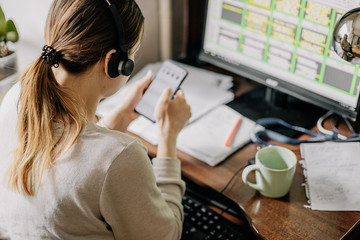 young woman working in an online call center arranged in her room by the window. Home environment, she drinks coffee, watches TV series on the phone and simultaneously works on computer