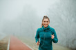 Beautiful fit girl athlete is running outside on foggy day. Girl is listening to music from her smartphone by using headset.