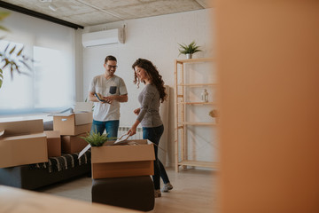 Photo of good looking young couple having fun while unpacking boxes in new home on moving day. Young couple is moving in to their new home.