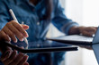 © NIKCOA - Young business woman hands holding electronic pen ( stylus pen ) and working on black digital tablet pc with laptop computer on the desk at modern home office. Remote working, online learning concept.