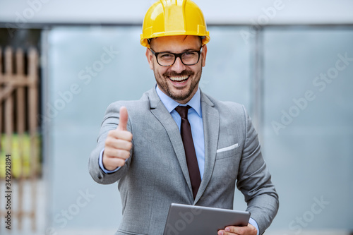 Attractive caucasian smiling elegant architect in suit, with eyeglasses and helmet on head holding tablet while standing at construction site and giving thumbs up Poster Mural XXL