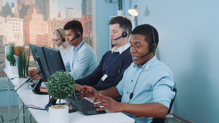 Wall Mural - Medium long shot of Black customer support agent working in busy call center by talking to the international client. His multiethnic team also taking calls.