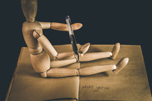 Wooden Figurine Holding Pen While Sitting On Open Book Against Black Background