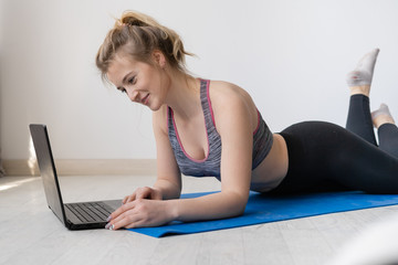 Pretty smiling young woman lying on the floor in sportswear in the living room and checking her social media on a laptop.