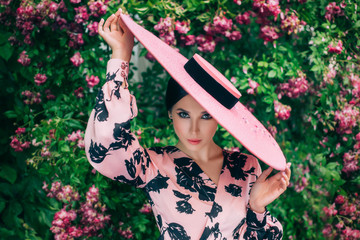 Wall Mural - Artwork portrait young happy charming chic woman. Vintage stylish elegant retro dress. Brunette enjoy spring holiday on March 8, beautiful nature blooming pink flowers tree, rose bush. Hat wide brim