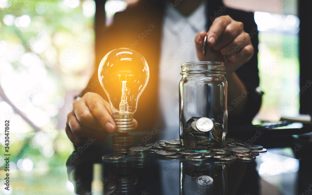Businesswoman holding a light bulb over coins stack on the table while putting coin into a glass jar for saving energy and money concept - obrazy, fototapety, plakaty 