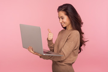 Wall Mural - Portrait of pleased young woman in casual sweater holding laptop and showing thumbs up while talking on video call, having online conference using computer. studio shot isolated on pink background