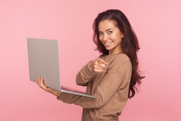 Wall Mural - Portrait of optimistic young woman in casual sweater holding laptop, pointing finger to camera and smiling, choosing you, freelance job advertisement. indoor studio shot isolated on pink background