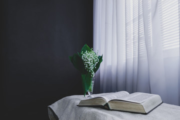 Wall Mural - Open Holy Bible and flower in vase on a white table, black   wall and window  background. Stay at home. Religion concept.