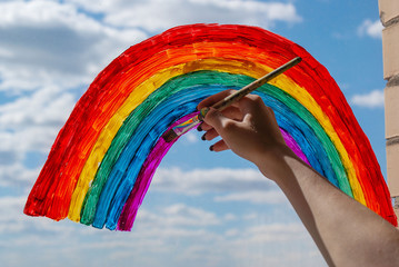 a young girl draws a rainbow on the window glass with a brush and gouache against a blue sky with white clouds. everything will be alright. symbol of happiness