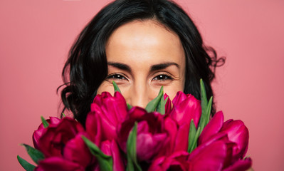 The 8th of March. Close-up photo of a cheerful brunette woman, who is looking in the camera and hiding a part of her face over the bunch of tulips.