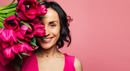 Girls' heaven. Portrait of a lovely brunette girl in a pink dress and golden necklace, who is looking in the camera, covering a part of her face with tulips.