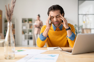 Wall Mural - Puzzled young man in yellow sweater sitting at desk with notepad and holding head in hands while suffering from headache at work