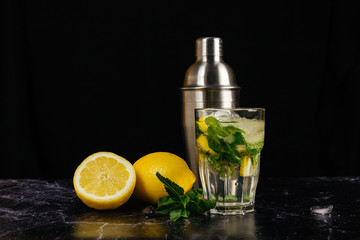 Fresh bright summer cocktail. Mojito cocktail with lemon, mint in a glass, metal cocktail shaker on a fashionable stone black background. Lemon mojito. 2 lemons, sprig of mint and a glass