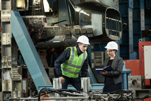 Portrait Of A Female Factory Manager In A White Hard Hat And Business Suit And Factory Engineer In Work Clothes. Controlling The Work Process At The Airplane Manufacturer. 