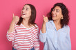 Closeup portrait of two happy smiling women wearing casual outfits, standing against pink studio background and pointing aside with their thumbs, ladies looking ti side with positive expression.